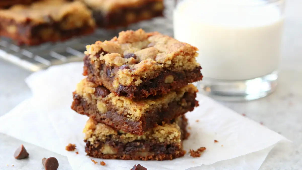 Stack of gooey brookies with layers of chocolate brownie and chocolate chip cookie, served with a glass of milk in the background.