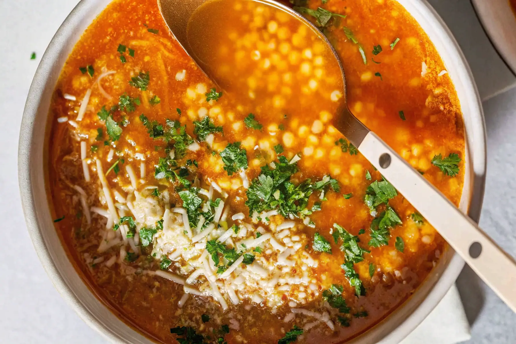 A close-up of a bowl of Pastina Soup topped with fresh parsley and grated Parmesan cheese, with a spoon dipped into the soup.